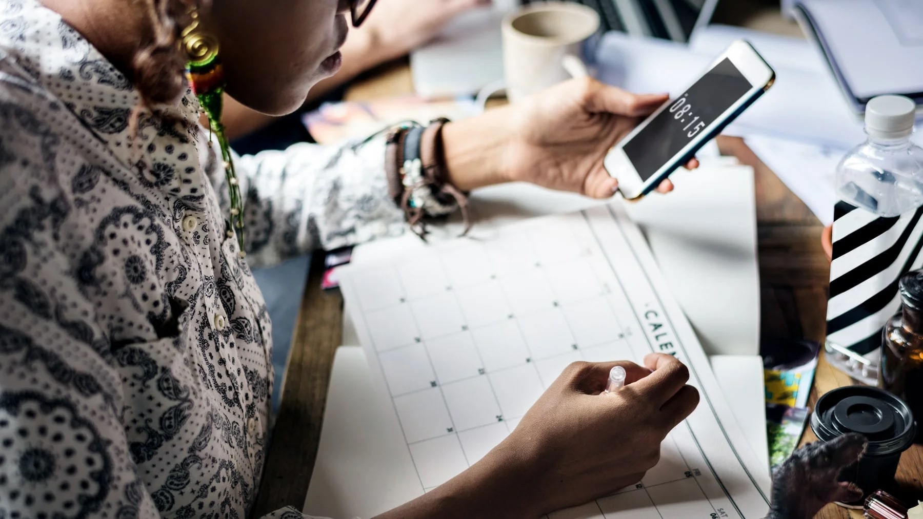Mulher marcando datas num calendário de papel sobre a mesa, com o celular na outra mão