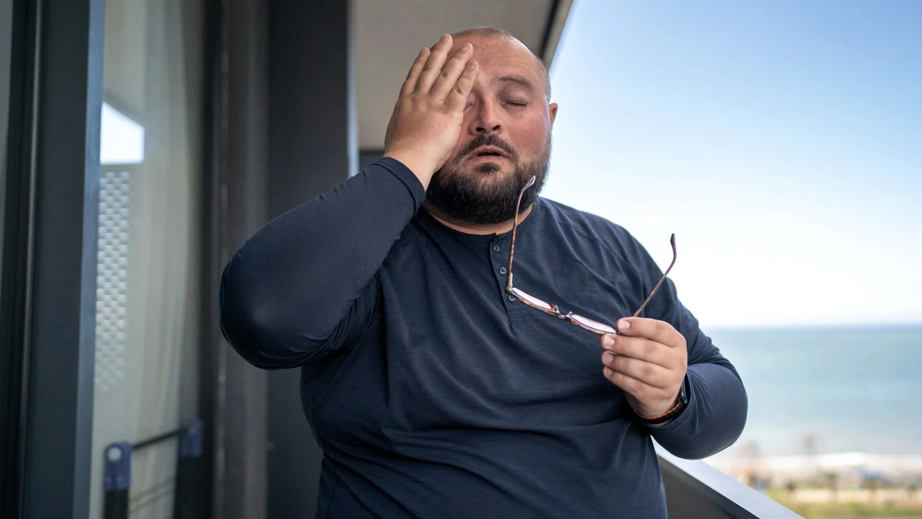 Homem cansado com a mão na testa na varanda, num momento de peso emocional.