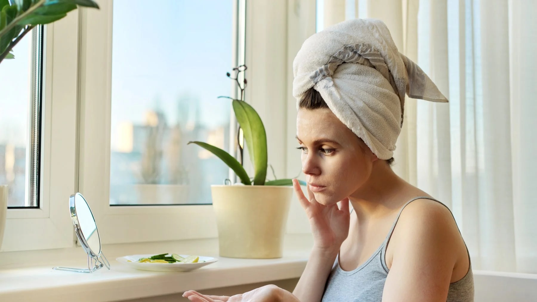 Mulher com toalha no cabelo passa creme no rosto perto da janela, com expressão cansada.
