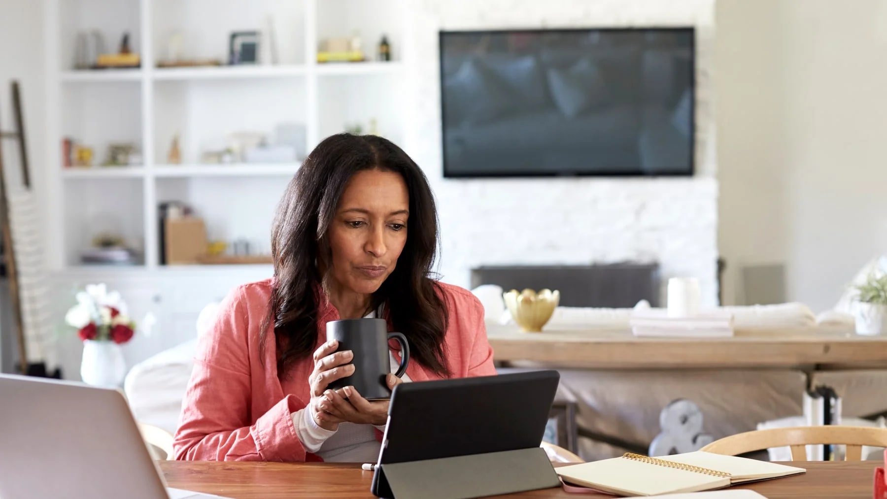 Mulher trabalhando em casa com notebook e tablet ao mesmo tempo, segurando uma caneca
