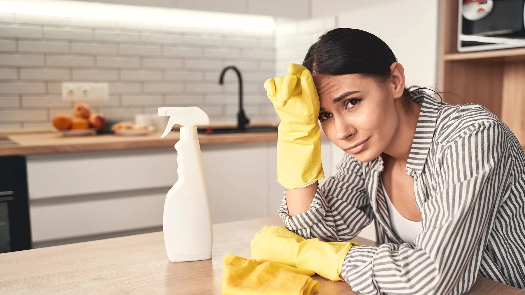 Mulher de luvas amarelas apoiada na bancada da cozinha, com a mão na testa, depois de limpar a casa.