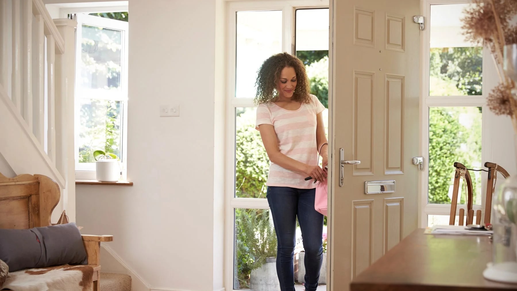 Mulher na porta de entrada de casa, com a bolsa na mão e a luz da manhã entrando pelo vão
