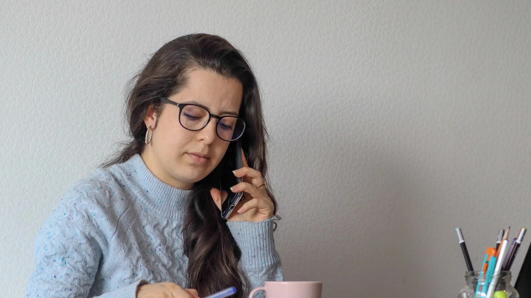 Mulher de óculos fala ao telefone e anota num caderno, sentada à mesa de trabalho em casa.