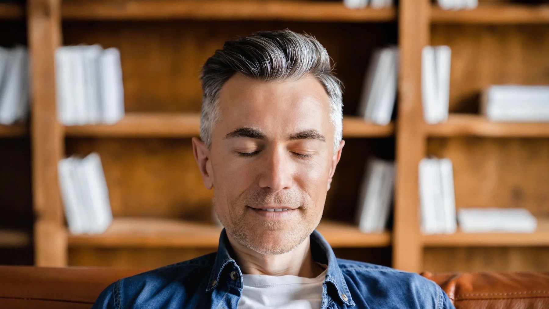 Homem de cabelo grisalho sentado no sofá, de olhos fechados e rosto tranquilo, com uma estante de livros atrás.