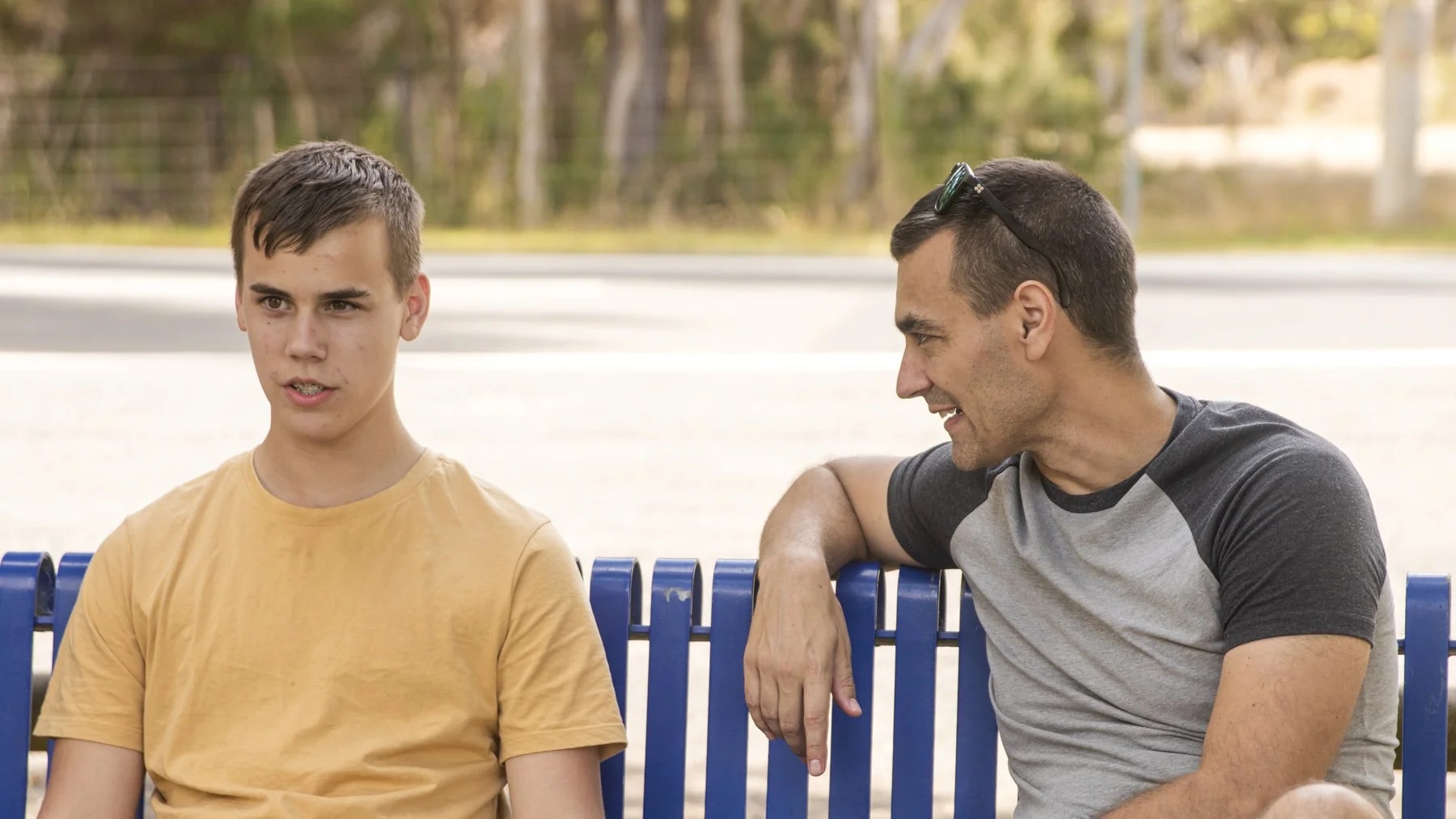 Pai e filho adolescente conversam sentados num banco, ao ar livre.