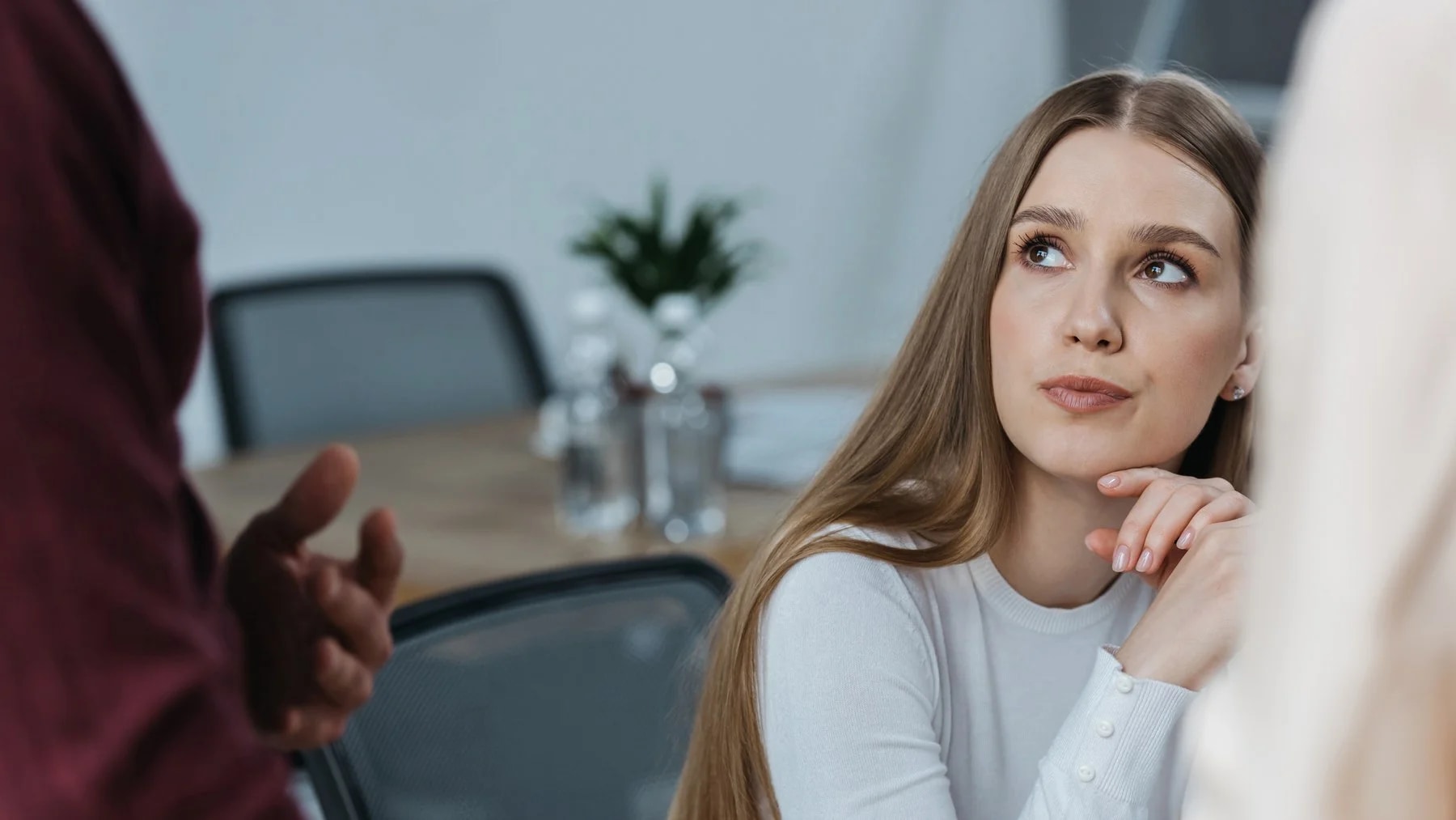 Jovem sentada numa reunião olha para cima, para alguém em pé que fala com ela, a mão no queixo.