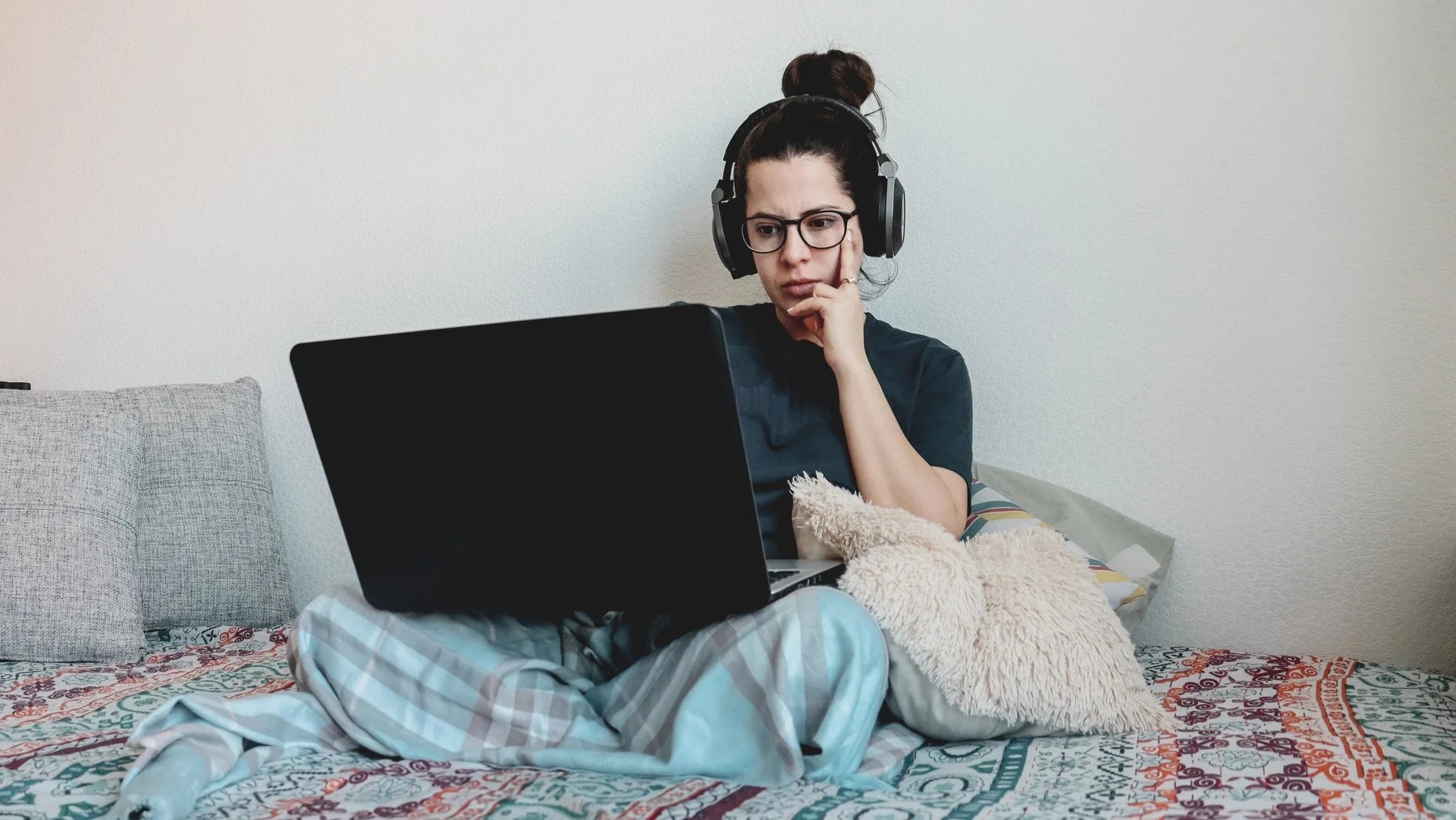 Jovem de fones de ouvido sentada na cama com o notebook no colo, a mão no queixo, pensativa.