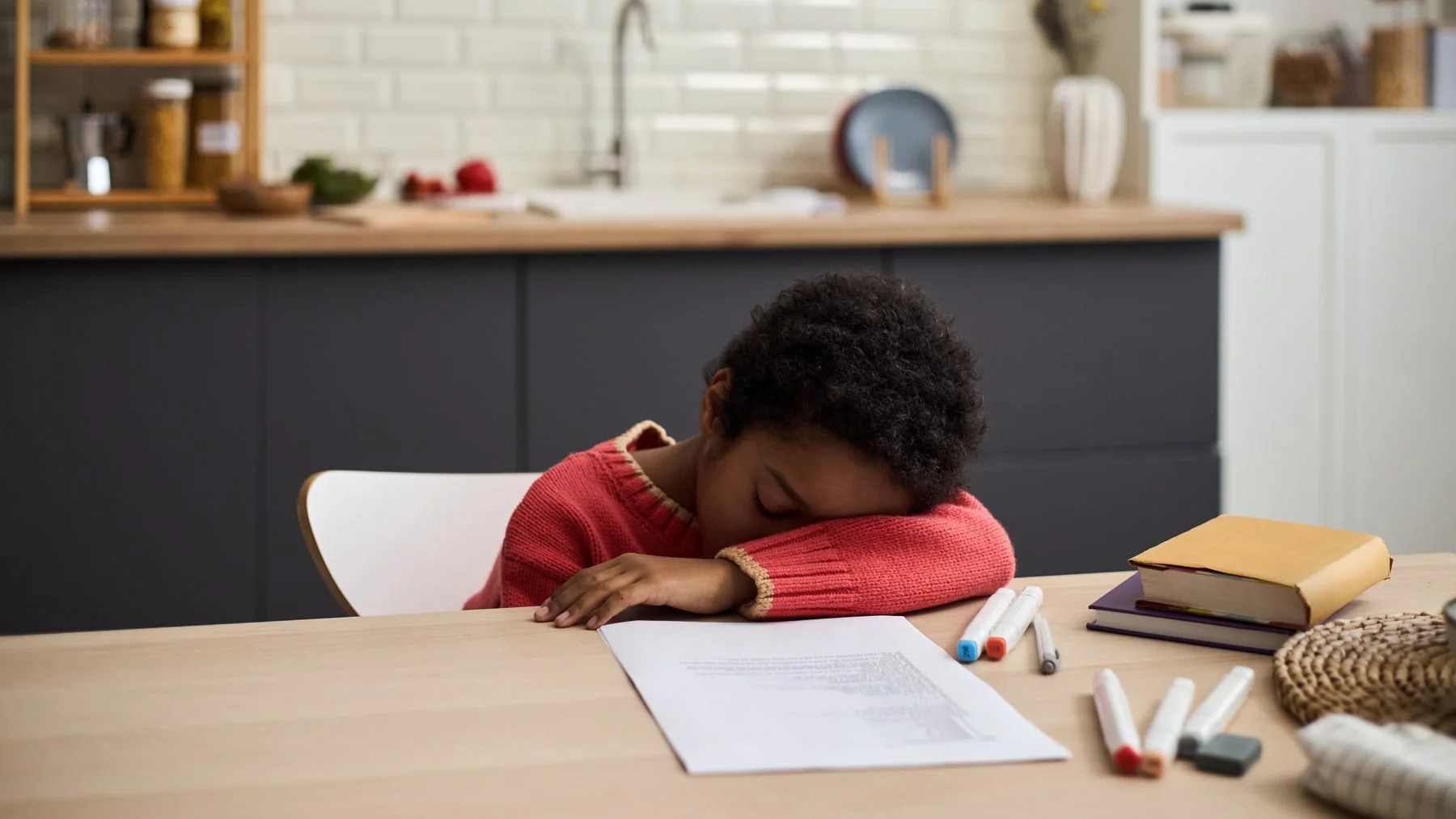 Criança com a cabeça apoiada na mesa da cozinha, ao lado do caderno e das canetas, cansada depois da lição