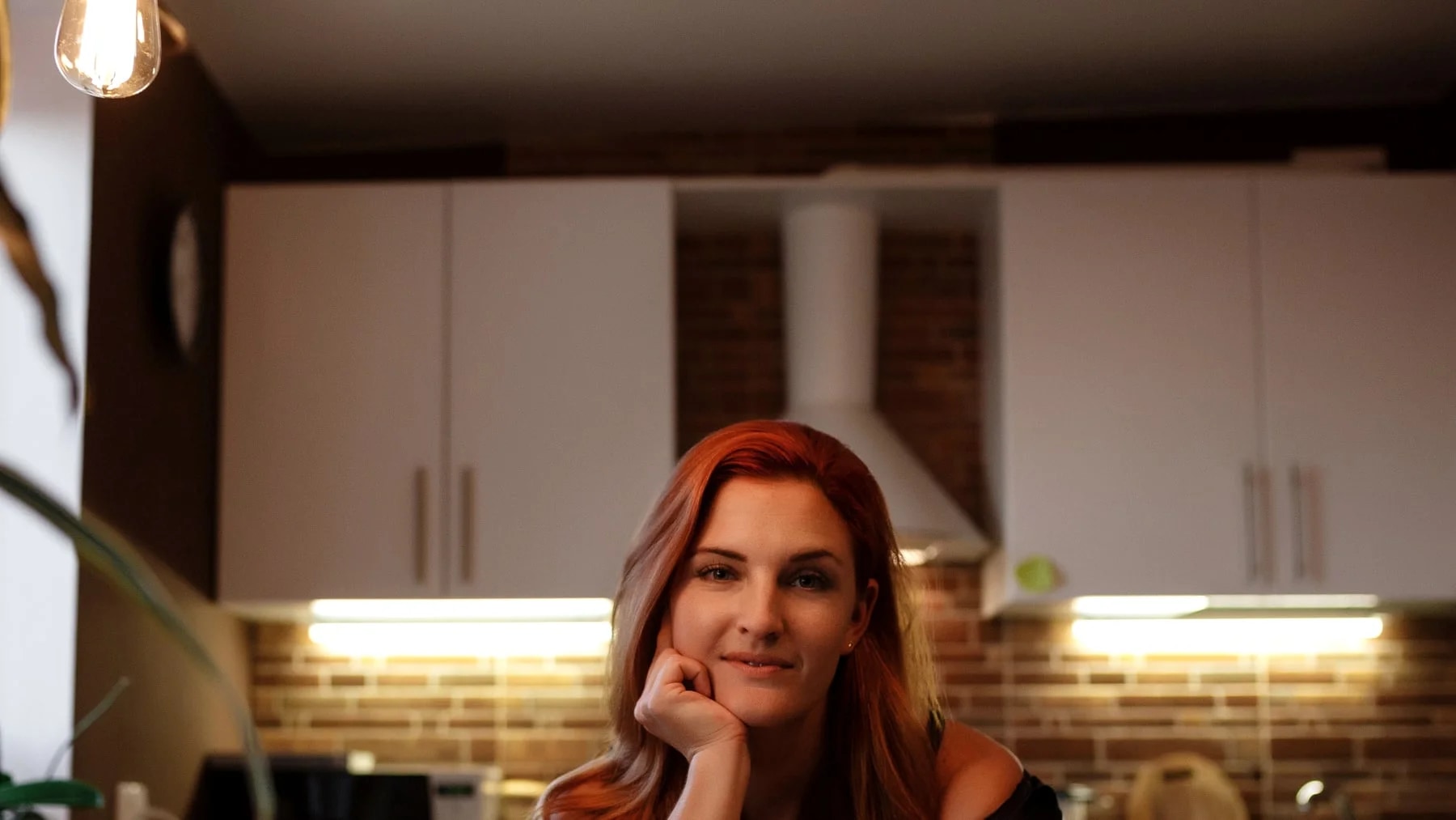 Mulher sozinha na cozinha de casa no fim do dia, apoiada na bancada, com a luz baixa das luminárias