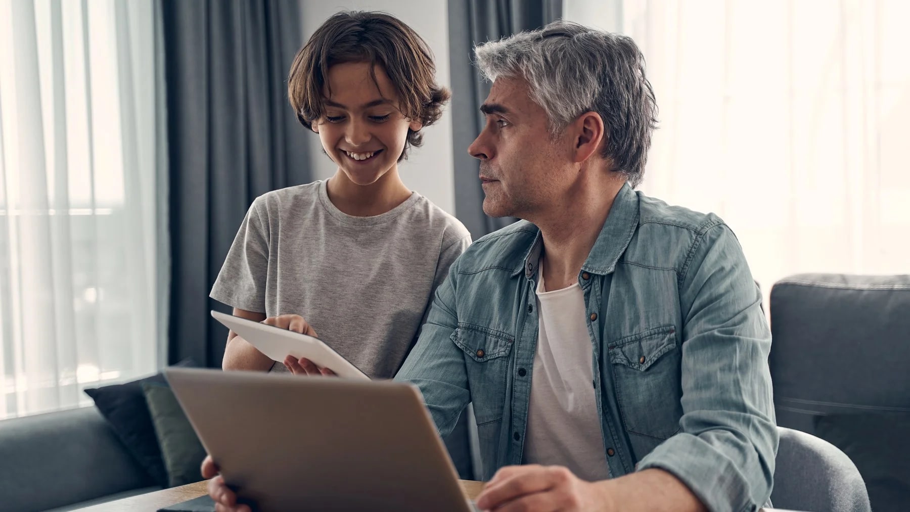 Pai de cabelo grisalho e filho adolescente sentados à mesa, olhando juntos a tela de um notebook.