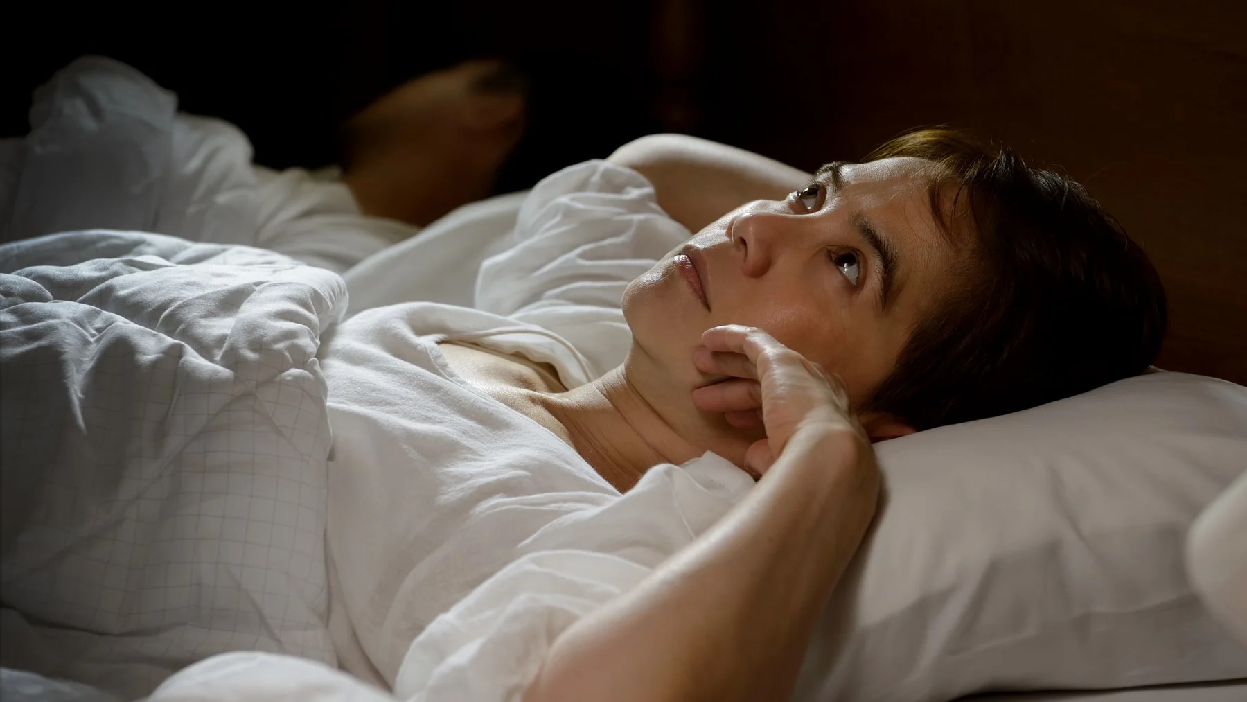 Mulher deitada na cama, acordada, olha para o teto com a mão perto do rosto.