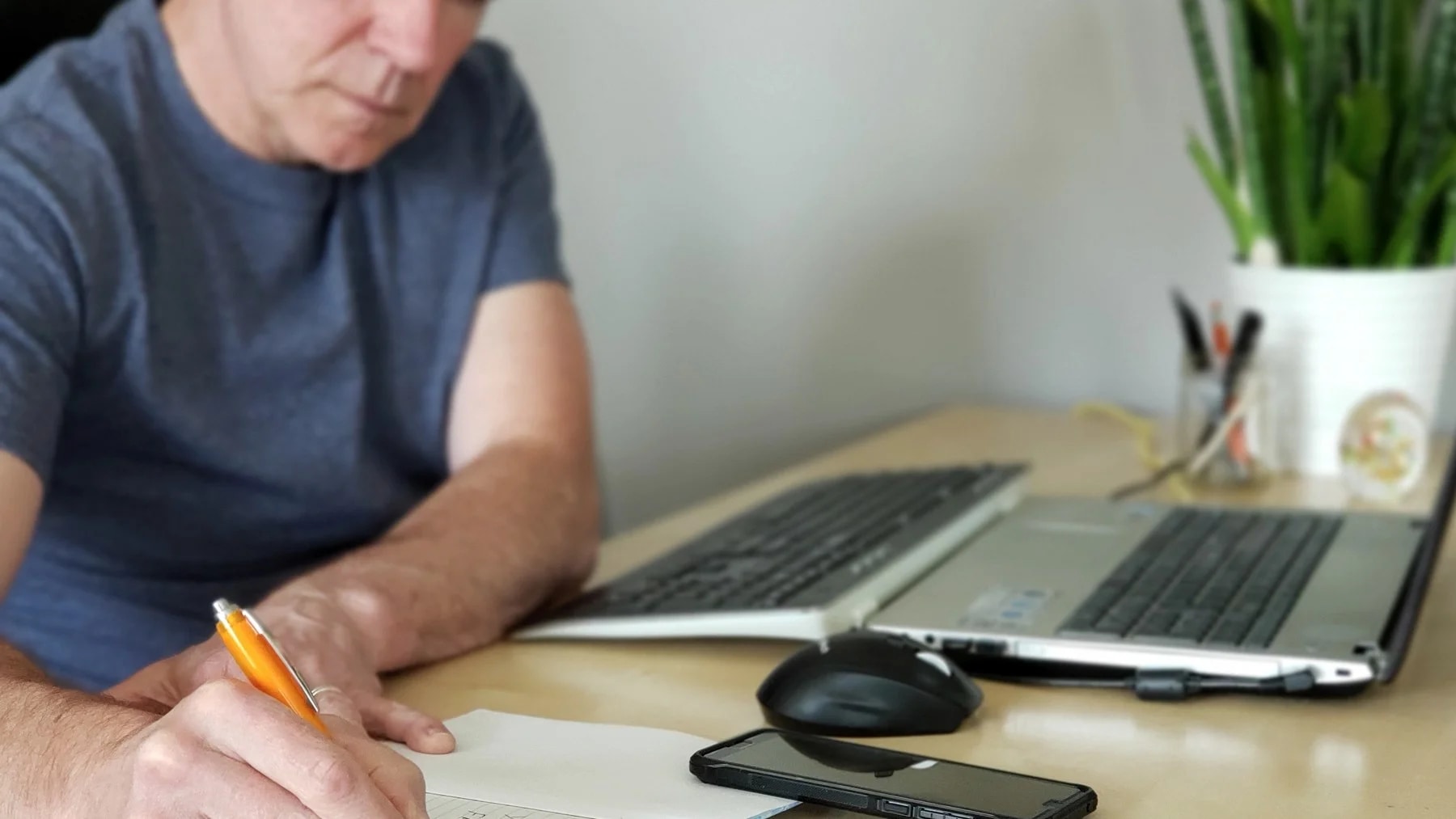 Homem anota num caderno, sentado à mesa com dois notebooks abertos e o celular ao lado.